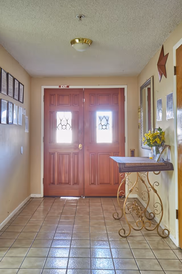 Interior hallway with a double wooden door featuring decorative glass panels and an exit sign above. The floor is tiled, and there is a decorative metal table with a cloth and a vase of yellow flowers on the right side. The walls have framed pictures and a mirror, with a star decoration on the right wall.