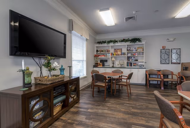 A common area in a senior living facility featuring a wall-mounted flat screen TV above a wooden cabinet with decorative items. There are several tables with chairs arranged around the room, a large window with blinds, and a white built-in shelving unit filled with books, decorations, and plants. The floor is wood, and the ceiling has recessed lighting.