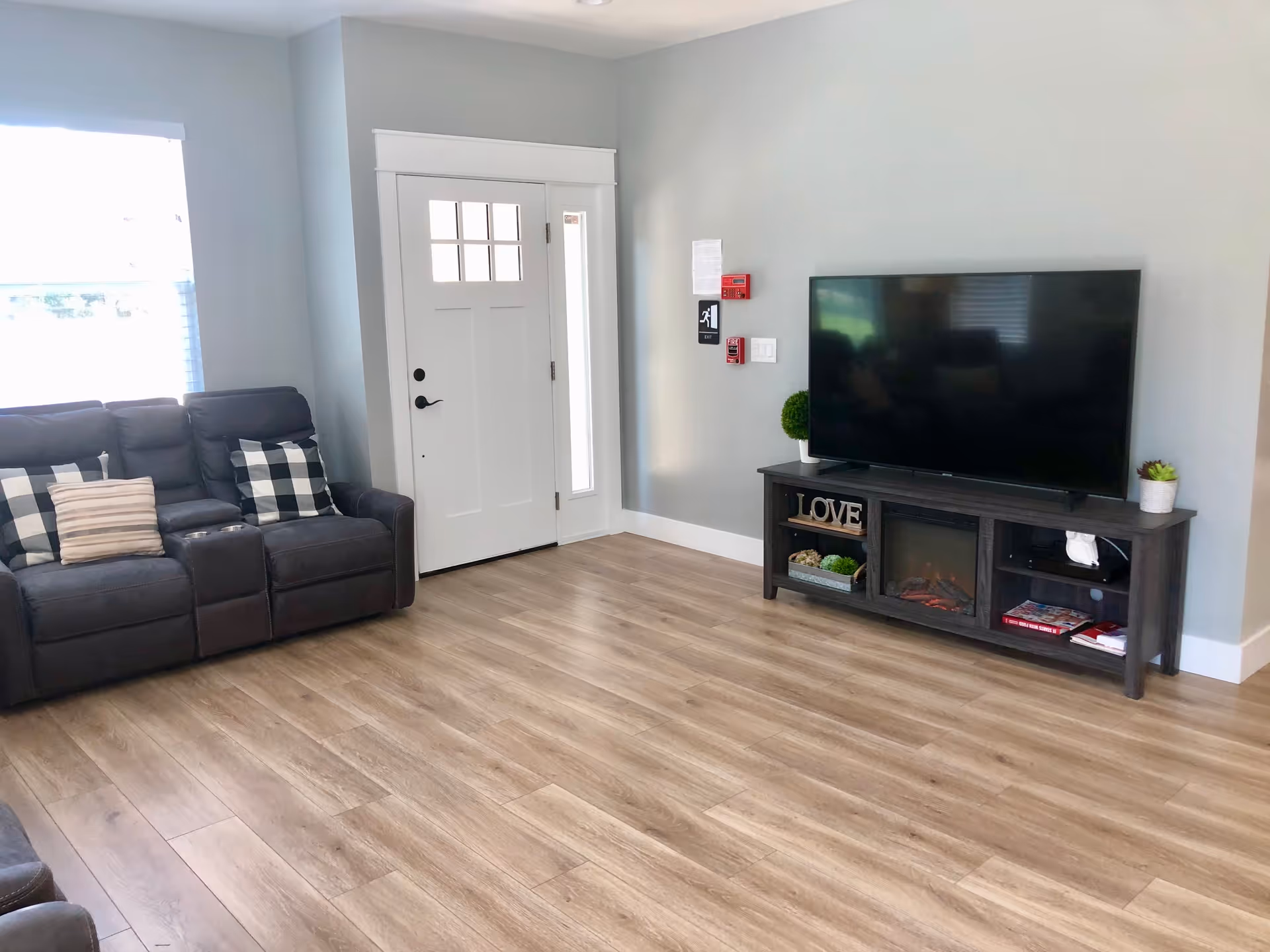 Bright living room with a reclining sofa, front door, large TV on a media console, and wood-look flooring.