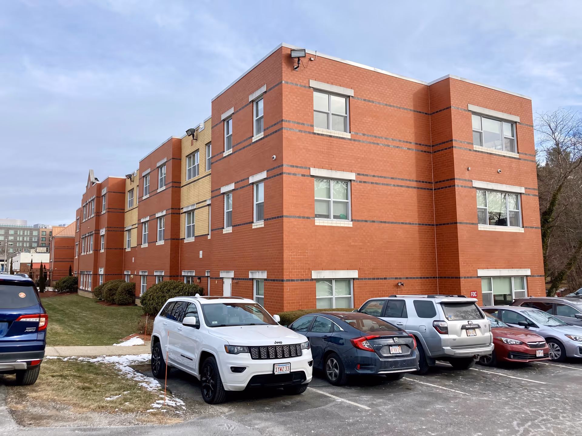 Three-story red-brick nursing facility with multiple parked cars in the front parking lot under a cloudy sky.