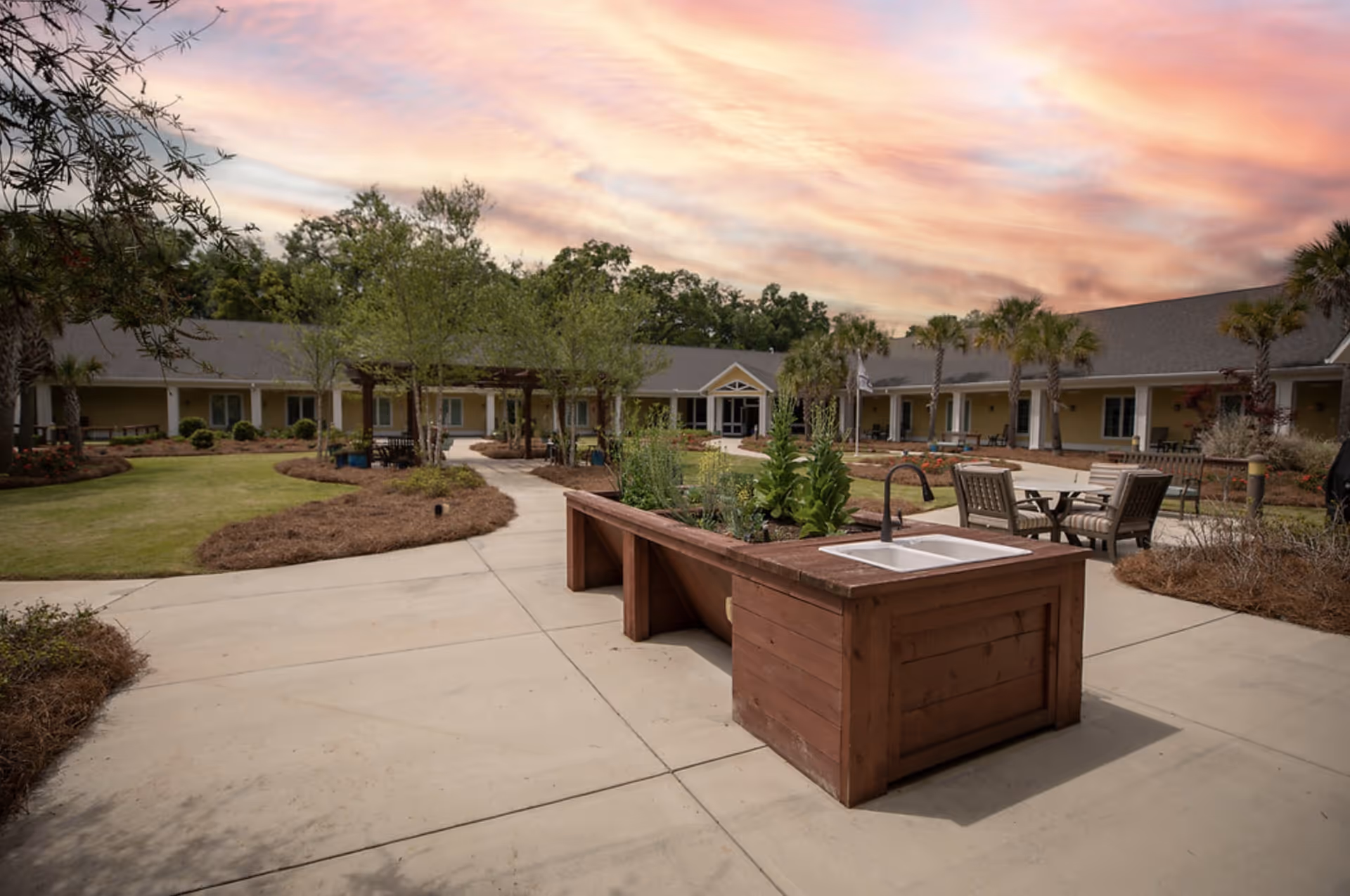 Outdoor courtyard area of a senior living facility with a wooden planter box featuring a built-in sink and faucet. The courtyard has paved walkways, green lawns, trees, and seating areas with tables and chairs. The building surrounds the courtyard with a covered walkway and multiple windows. The sky is colorful with a sunset or sunrise.