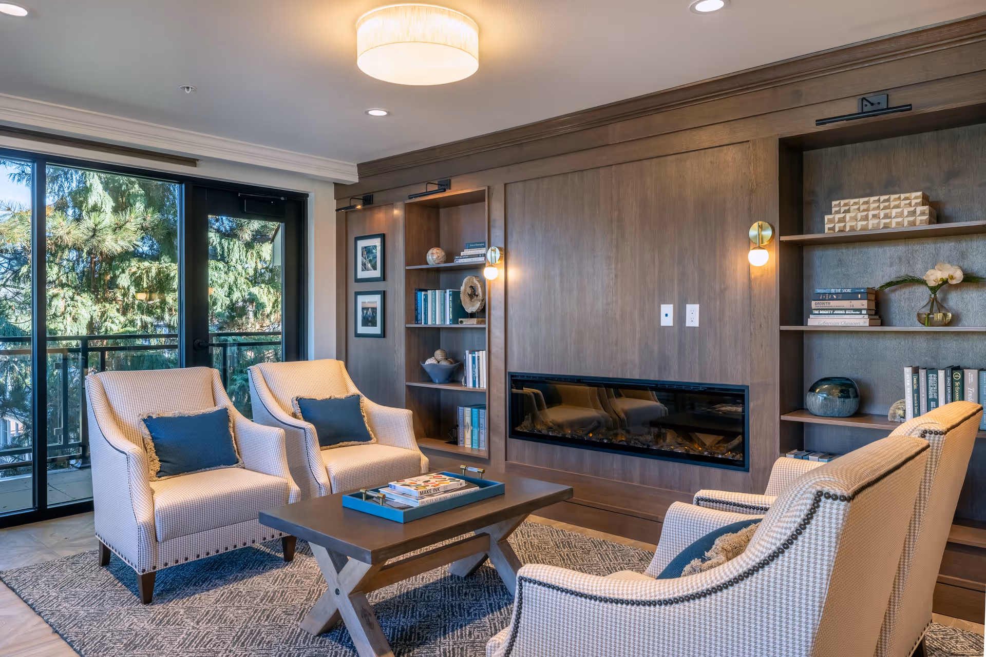 A cozy senior living room with four beige armchairs arranged around a wooden coffee table. The room features a modern electric fireplace built into a wooden wall with built-in shelves holding books and decorative items. Large glass doors open to a balcony with a view of green trees outside. The room is softly lit by a ceiling light and wall sconces.