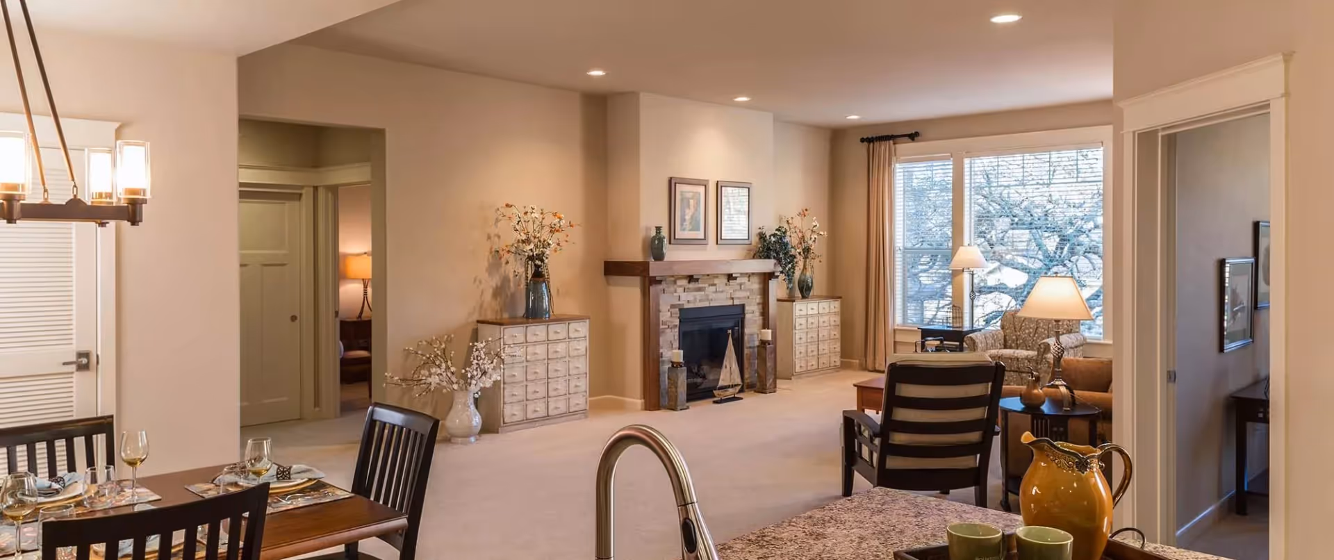 A cozy and well-lit living room and dining area in a senior living facility. The living room features a stone fireplace with a wooden mantel, two decorative cabinets with vases and flowers, a large window with beige curtains, and comfortable seating including armchairs and a side table with a lamp. The dining area has a wooden table set with placemats, wine glasses, and napkins, with a modern chandelier overhead. The foreground shows a kitchen counter with a faucet, a yellow pitcher, and green mugs.