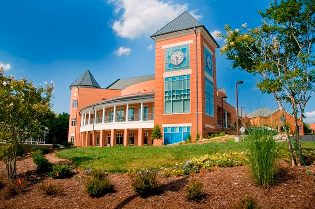 Brick senior living building with a prominent clock tower, curved balcony, and landscaped lawn under a blue sky.