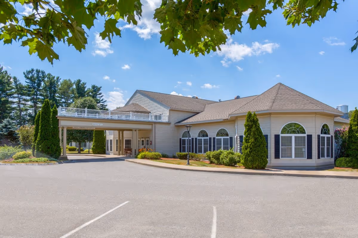 Exterior view of Benchmark at Haverhill Crossings senior living facility on a sunny day, showing a large building with beige siding, multiple arched windows with black shutters, a covered entrance supported by columns, landscaped greenery including bushes and tall trees, and a clear blue sky with some clouds.