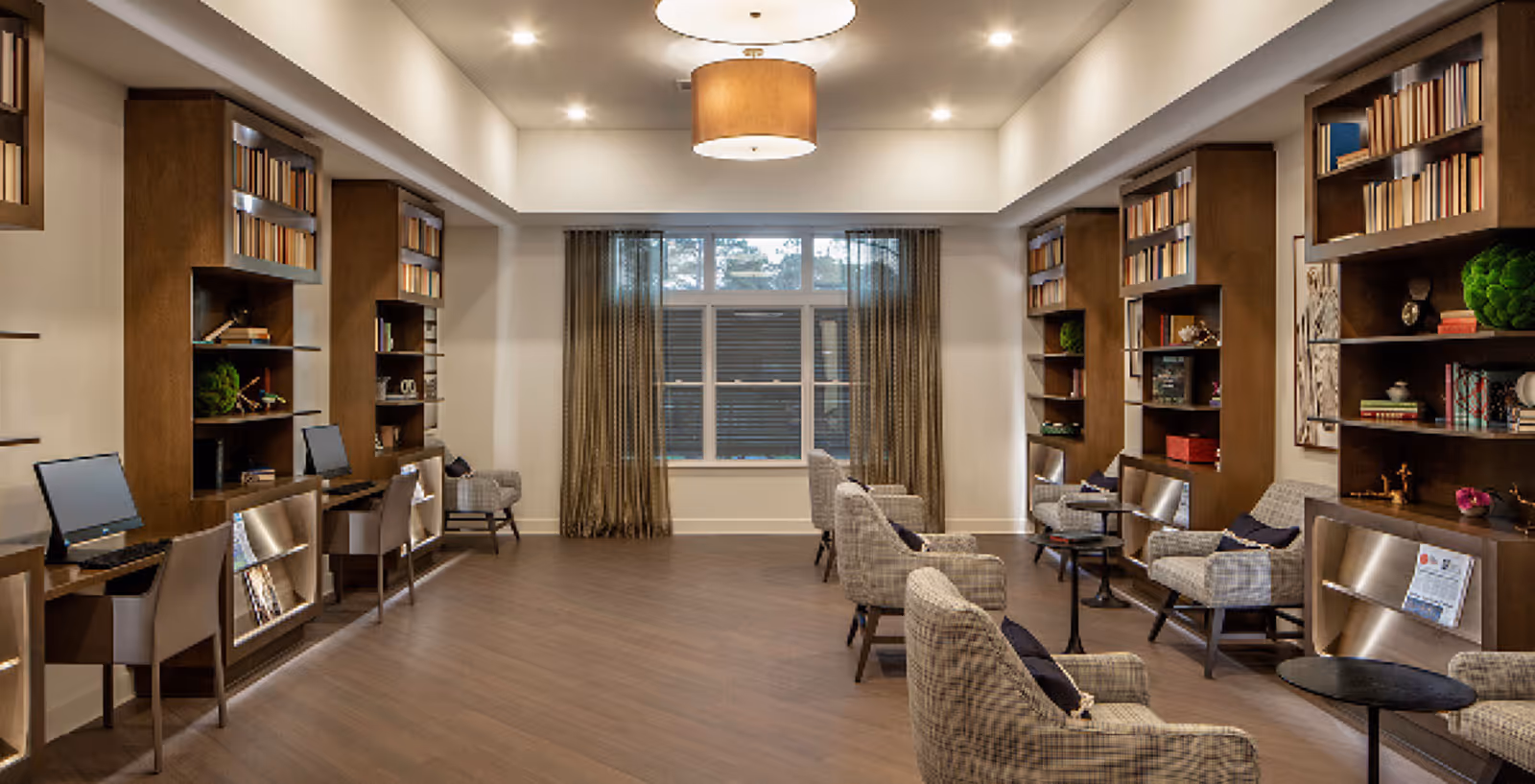 A spacious, well-lit common area with wooden bookshelves filled with books and decorative items on both sides. There are several comfortable armchairs with cushions arranged facing each other and small round tables between them. On the left side, there are desks with computers and chairs. A large window with curtains is at the far end of the room, and a modern ceiling light fixture hangs above.