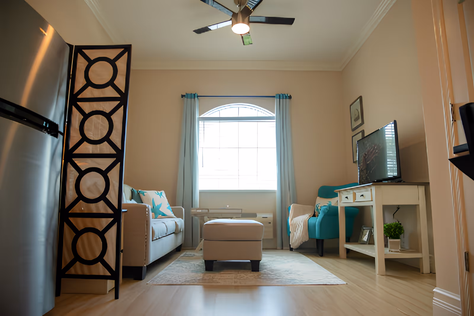 A cozy living room with a beige sofa adorned with starfish-patterned pillows on the left, a teal armchair with a throw blanket on the right, and a beige ottoman in the center on a light-colored rug. A white wooden TV stand holds a flat-screen TV and a small potted plant. A large window with light blue curtains lets in natural light, and a ceiling fan with a light fixture is mounted above.