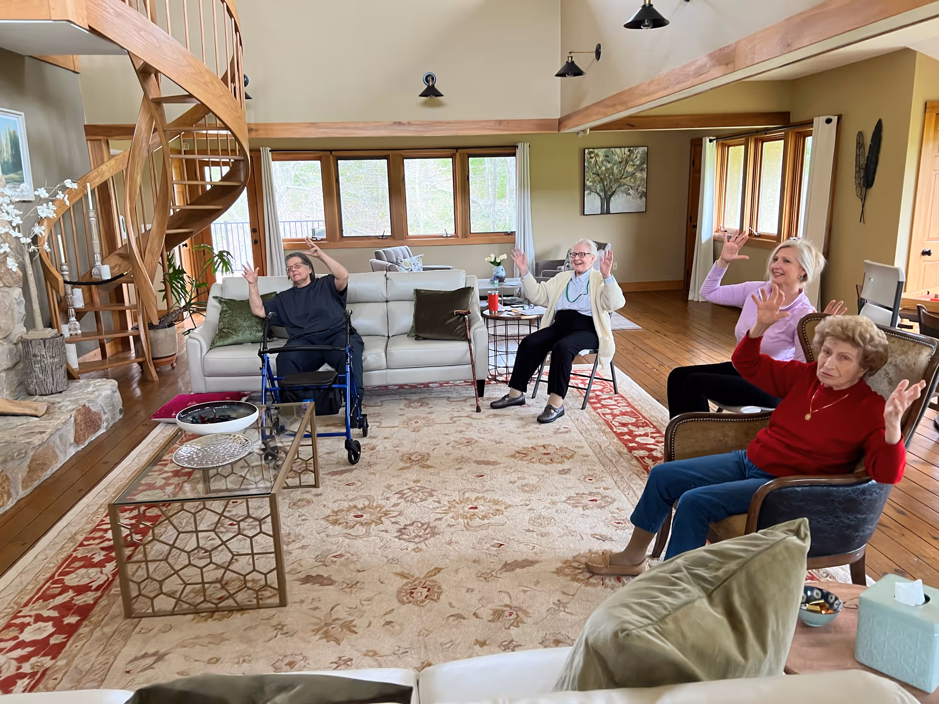 A spacious living room with wooden floors and a large patterned area rug. Four elderly women are seated in chairs and a sofa, smiling and raising their hands. The room features a spiral wooden staircase, large windows with natural light, a stone fireplace, and various pieces of furniture including a glass coffee table and side tables with decorative items.