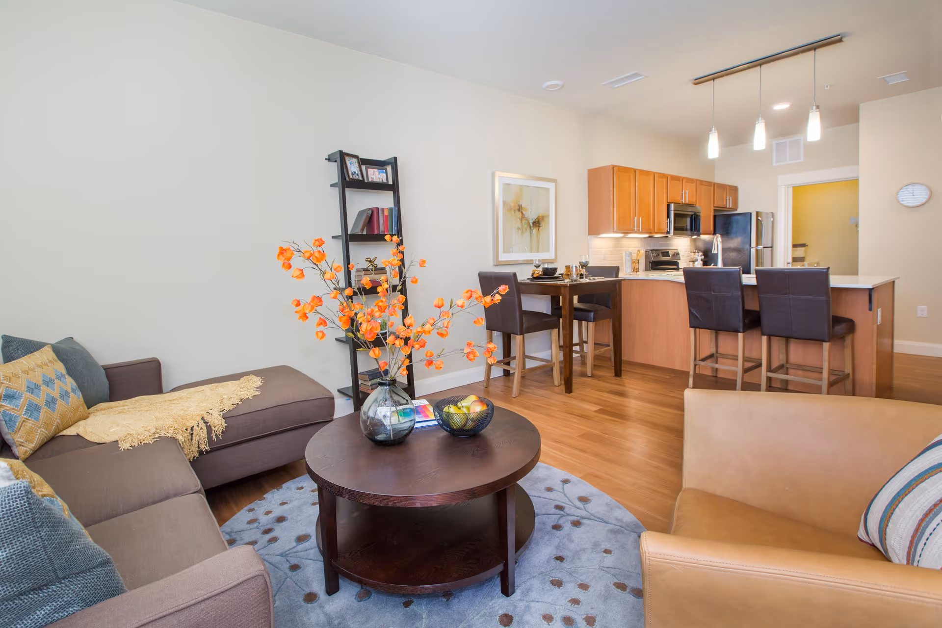 A bright and modern open-concept living area featuring a brown sectional sofa with colorful pillows and a yellow throw, a round dark wood coffee table with a vase of orange flowers and a bowl of fruit, a tan armchair with a striped pillow, and a kitchen area with wooden cabinets, stainless steel appliances, a breakfast bar with two dark leather stools, and a small dining table with two chairs. A black ladder shelf with books and framed photos is against the wall, and a framed abstract painting hangs above the dining table.