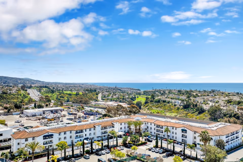 Aerial view of San Clemente Villas with a red-tile roofed apartment complex, nearby greenery and roads, and the ocean on the horizon.