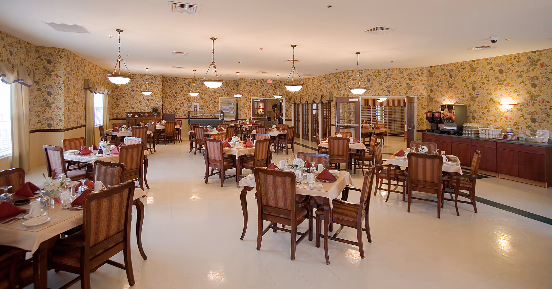 Spacious senior dining room with multiple set tables and wooden chairs, floral wallpaper, and a buffet/serving area.