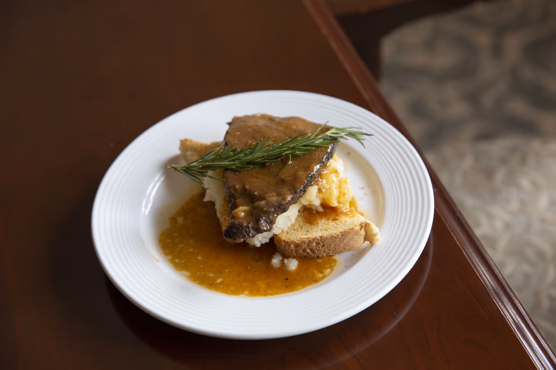 A white plate with a serving of meatloaf topped with gravy and a sprig of rosemary, placed on a slice of bread with mashed potatoes underneath, sitting on a wooden table.