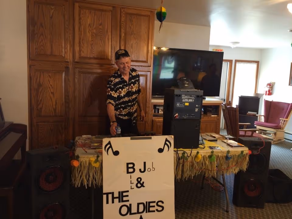 A man stands behind a decorated table with speakers and a sign reading 'B J & THE OLDIES' in a communal living room area with a large TV and wooden cabinets.