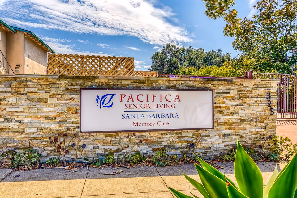 Stone wall with a sign that reads 'Pacifica Senior Living Santa Barbara Memory Care' under a partly cloudy blue sky, with some plants and trees around.