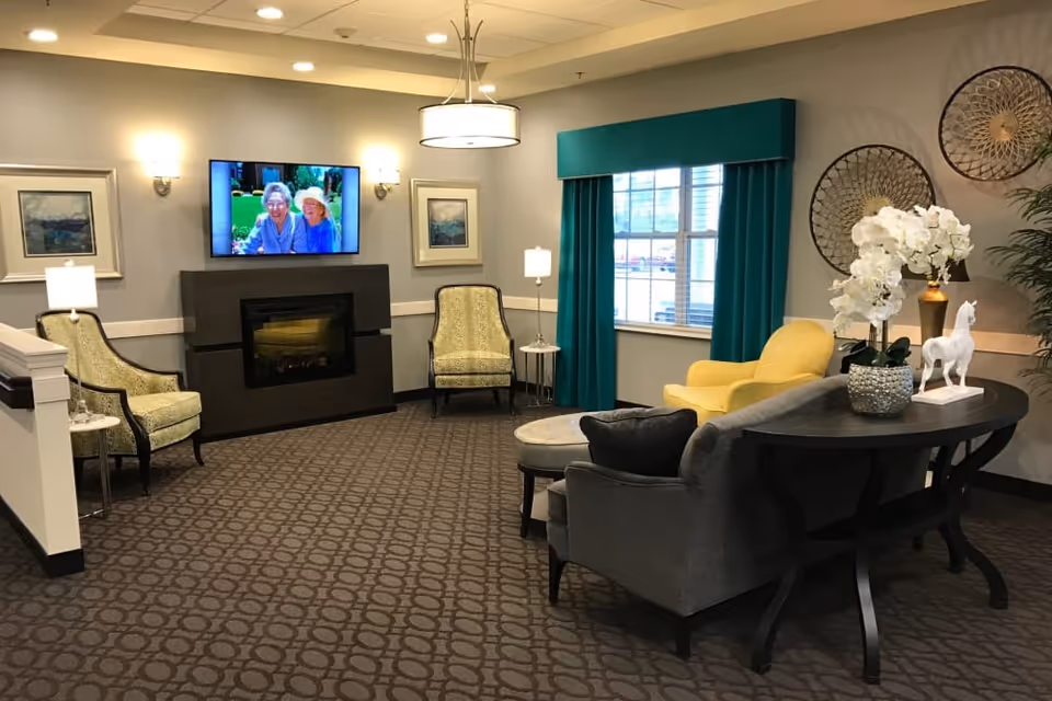 A cozy living room area in a senior living facility with patterned carpet, a dark wood fireplace with a TV mounted above it displaying two elderly women, two yellow patterned armchairs on either side of the fireplace, a large window with teal curtains, a yellow armchair, a gray sofa, and a dark wood console table decorated with white flowers and a white horse figurine.
