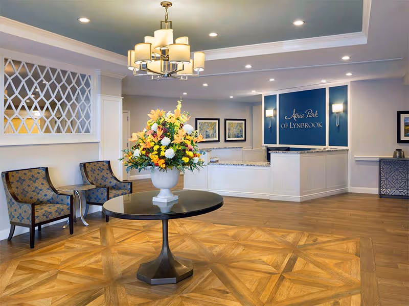 Reception area of Atria Park of Lynbrook featuring a round table with a large floral arrangement in the center, two patterned armchairs with a small table between them on the left, a chandelier hanging from the ceiling, and a reception desk with a blue sign that reads 'Atria Park of Lynbrook' in the background.