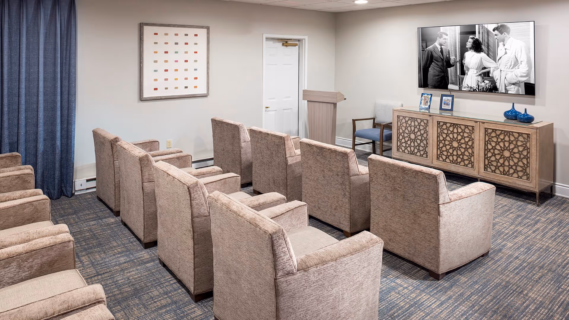 A small theater-style room with two rows of beige upholstered armchairs facing a wall-mounted flat screen TV showing a black and white movie. The room has a patterned carpet, a wooden cabinet with decorative front panels beneath the TV, a podium, and a single chair against the wall. A framed abstract artwork hangs on the wall beside a closed white door.