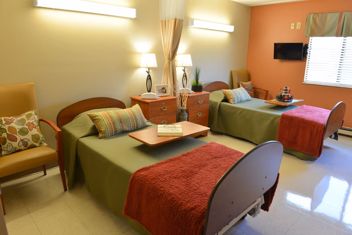 A bright and clean room with two single beds covered in green bedspreads and red throws. Each bed has a decorative pillow and a wooden overbed table. Between the beds is a wooden nightstand with two lamps, a photo frame, and a plant. There are two armchairs with patterned cushions, a window with blinds, and a small wall-mounted TV on an orange accent wall.