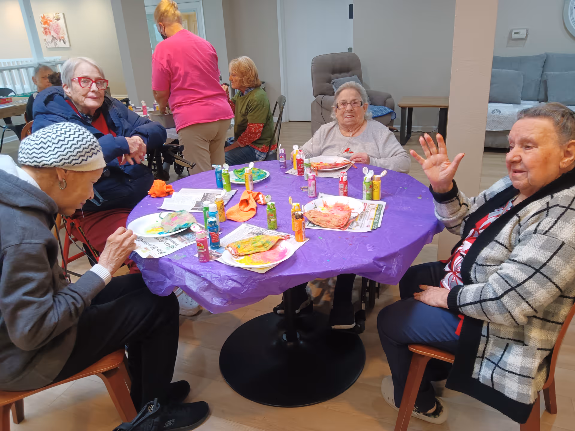 A group of elderly women sitting around a table covered with a purple tablecloth, engaged in a painting activity with various paint bottles and colorful artwork on paper plates. One woman is waving at the camera, and a staff member in a pink shirt is standing nearby. The room has a cozy, communal atmosphere with chairs and a couch in the background.