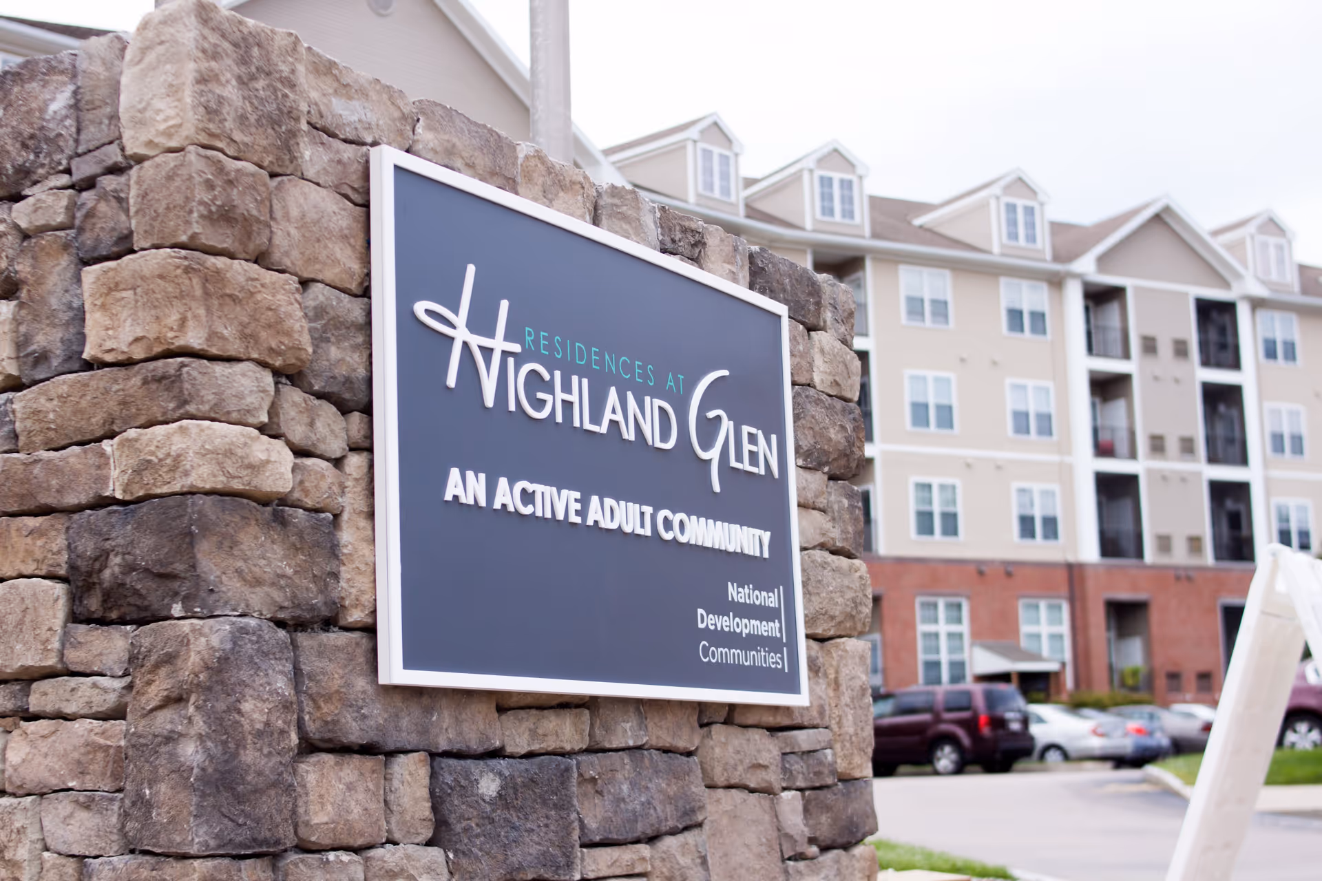 Stone monument sign for Residences at Highland Glen, an active adult community, with a multi-story residential building and parked cars in the background.
