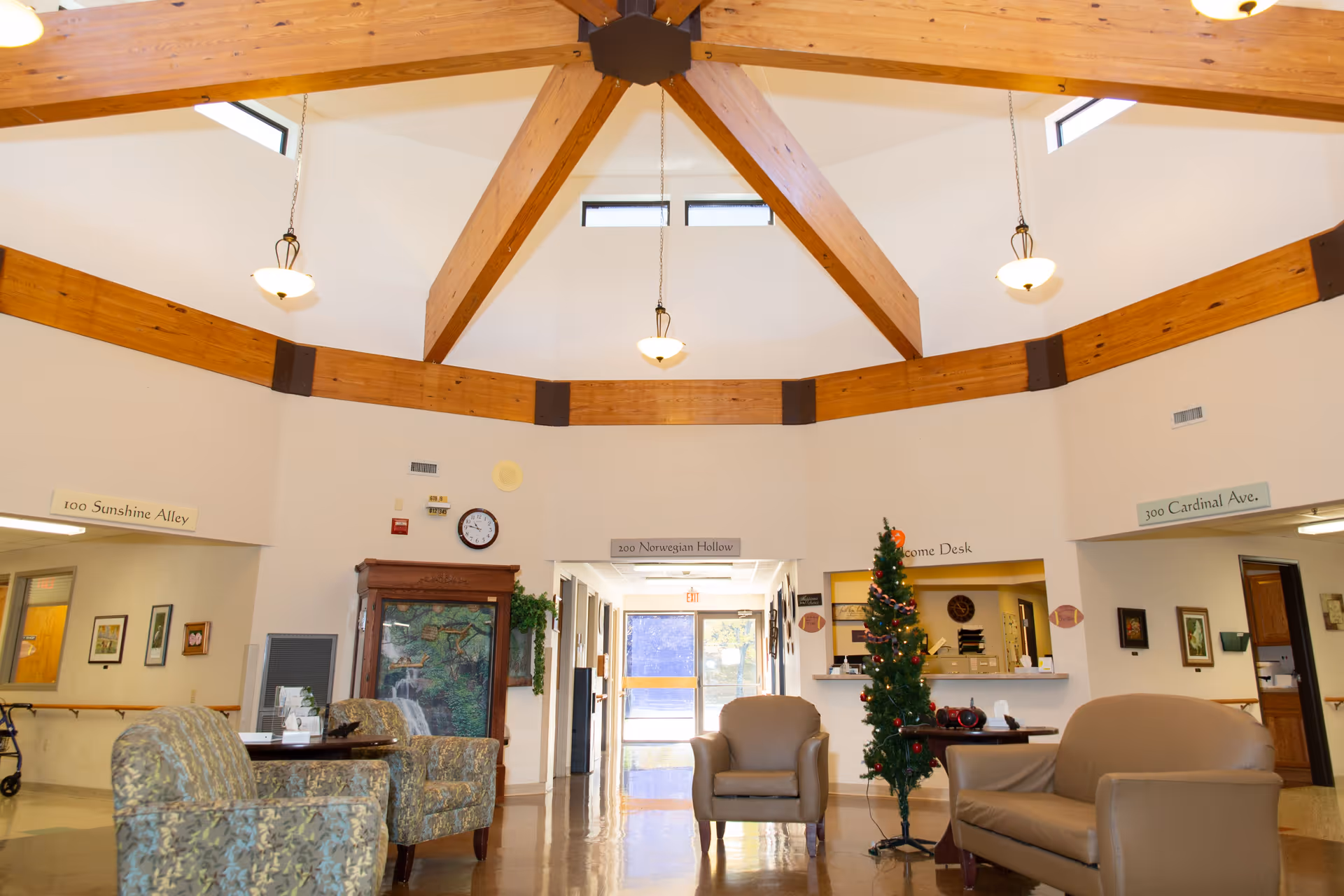 A spacious and well-lit common area in a senior living facility with wooden beams on the ceiling, several armchairs and a sofa arranged around a small table, a decorated Christmas tree, and signs indicating different hallways and a welcome desk.