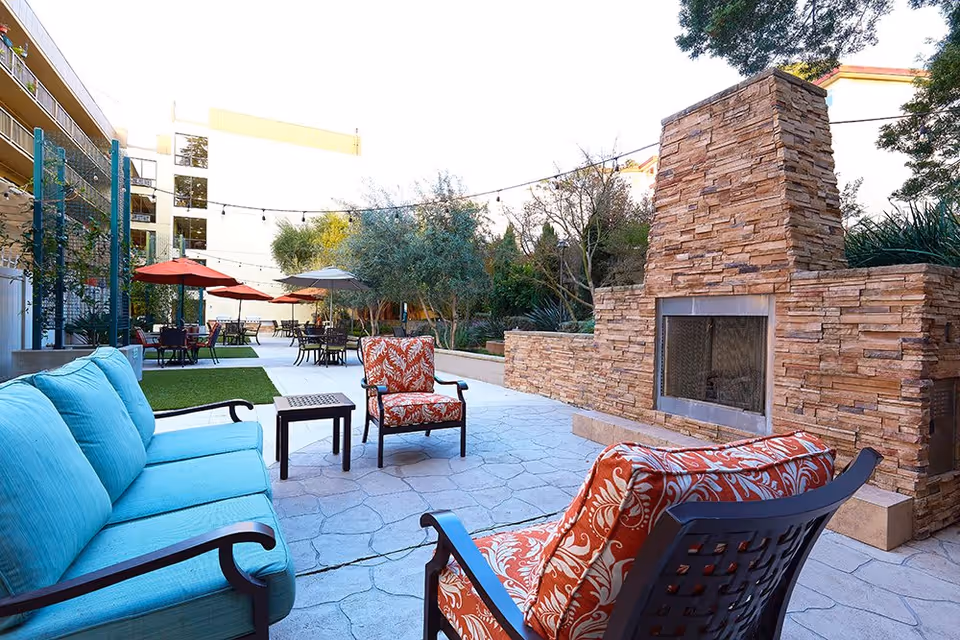 Outdoor patio area at The Point At Rockridge Senior Living featuring cushioned seating including a turquoise sofa and two red patterned armchairs around a stone fireplace. There are tables with umbrellas and string lights overhead, surrounded by trees and greenery.