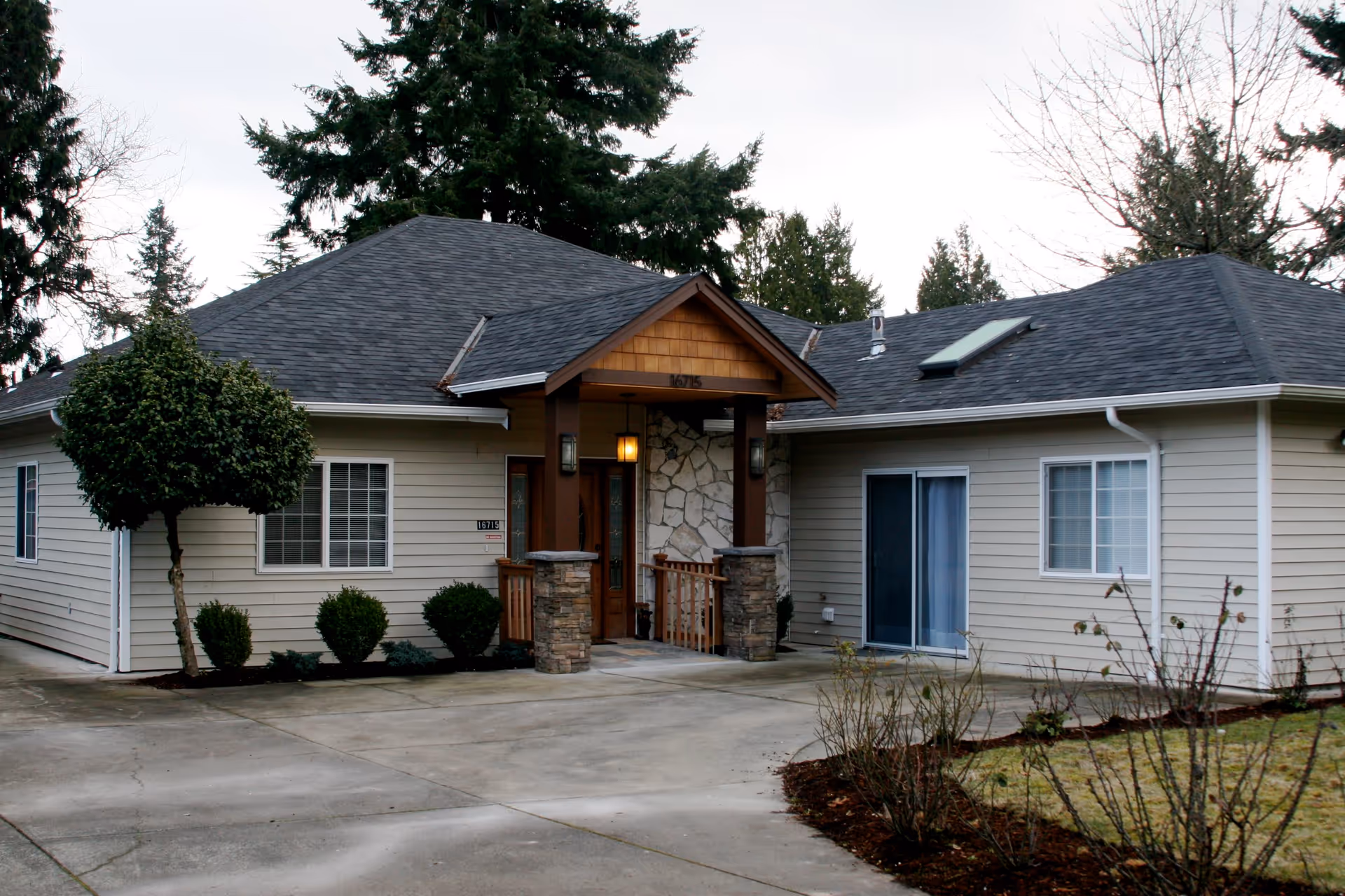 Exterior view of a single-story residential building with beige siding, a dark shingled roof, and a covered entrance supported by stone pillars. There are several windows, a sliding glass door, and a concrete driveway in front. Surrounding the building are some small bushes, a tree, and a lawn area with bare shrubs. Tall evergreen trees are visible in the background under a cloudy sky.
