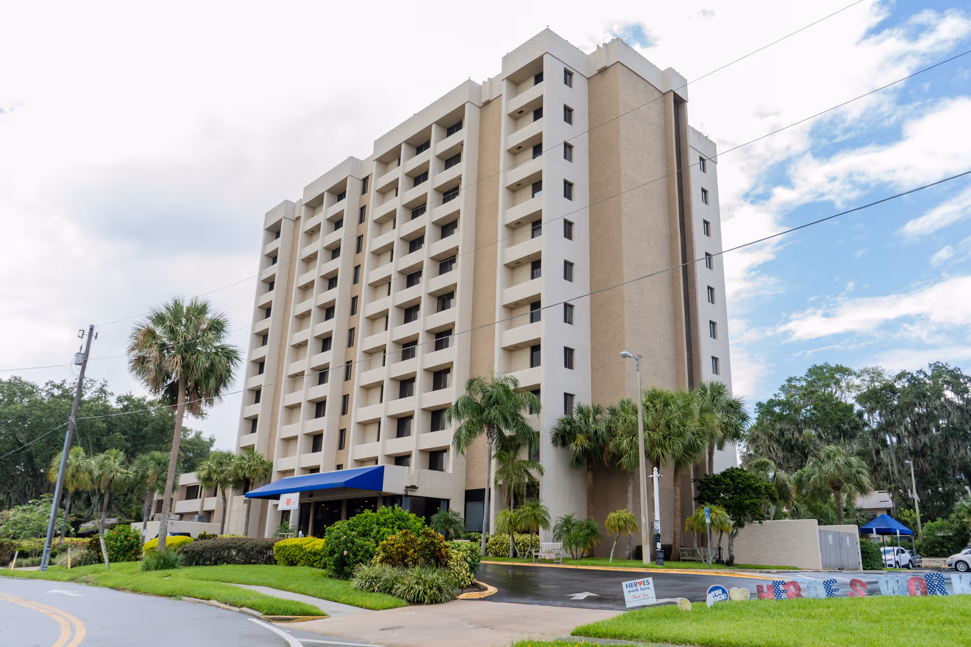 Exterior view of a multi-story beige senior living facility building with balconies, surrounded by palm trees and greenery under a partly cloudy sky.