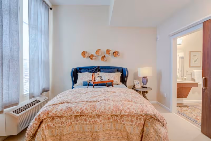 A cozy bedroom with a bed featuring a patterned comforter and a dark blue upholstered headboard. A wooden tray with a teapot and cups is placed on the bed. To the right is a small round side table with a lamp and a framed photo. The room has light-colored walls and a window with gray curtains on the left. An open doorway on the right leads to a bathroom with a sink, mirror, and towel rack.