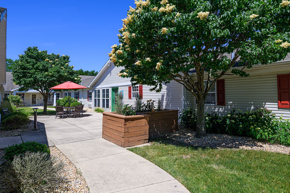 Outdoor courtyard area of a senior living facility with a concrete walkway, green grass, flowering trees, and a seating area with a table and chairs under a red umbrella. The building has white siding with red shutters and a green door.