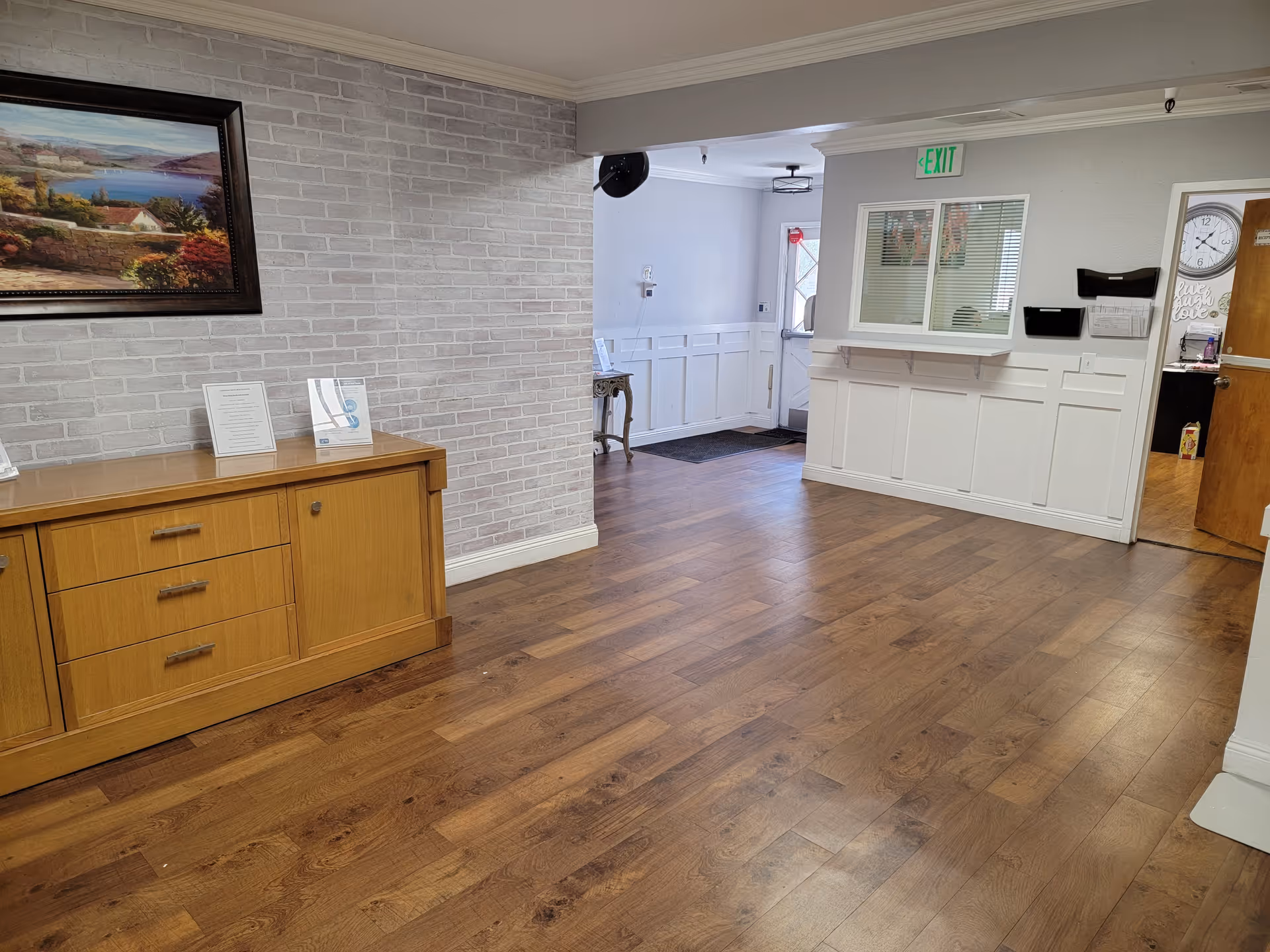 Interior view of a hallway or lobby area with wooden flooring, a wooden cabinet with drawers on the left, a framed landscape painting above the cabinet, white brick wall, and a reception window on the right. There is an exit door in the background with an illuminated green exit sign above it.