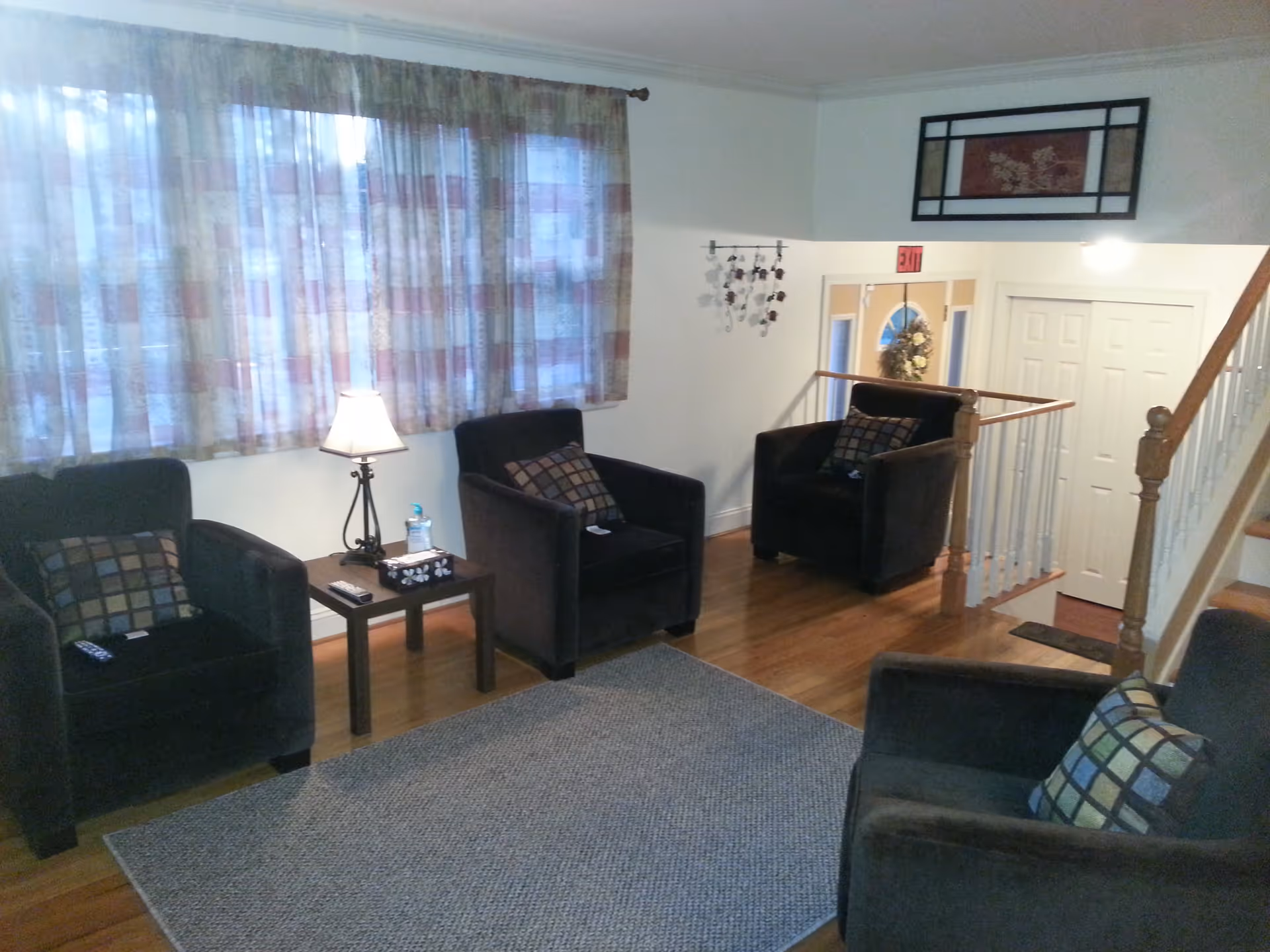 A cozy living room with four upholstered armchairs around a rug, a side table with a lamp, and a staircase and doorway in the background.