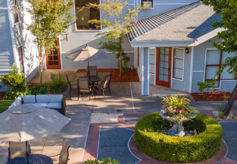 Outdoor courtyard area of a senior living facility with patio tables and chairs, umbrellas, a circular hedge with a decorative planter in the center, and surrounding trees and shrubs. The building exterior features light blue siding with red-framed windows and doors.