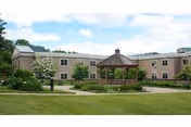 Two-story beige nursing facility building with a central wooden gazebo and landscaped lawn under a partly cloudy sky.
