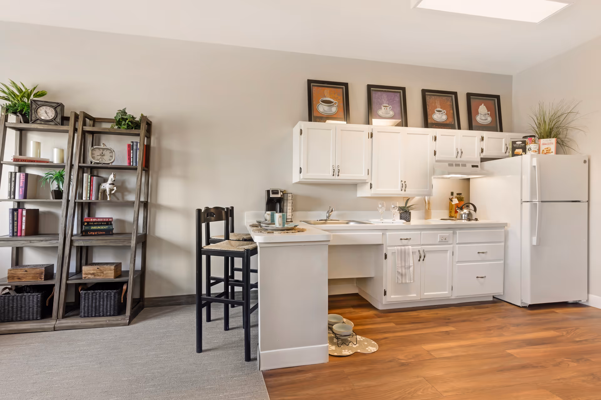 Bright kitchenette with white cabinets, a refrigerator, a small breakfast bar with stools, and a shelving unit against a light wall.