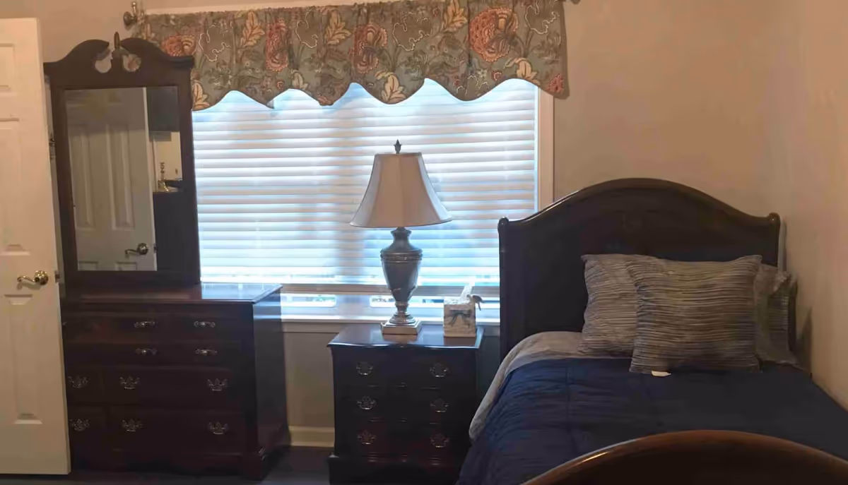 Small single bedroom with a bed, nightstand and dresser in front of a window with blinds and a patterned valance.