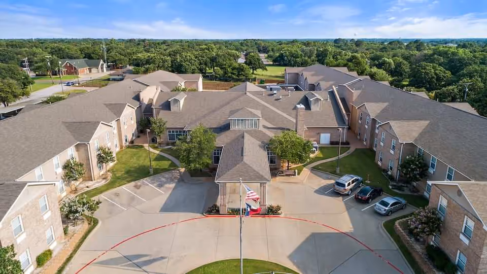 Aerial view of Morada Pantego senior living facility showing a large, multi-wing building with a central entrance, surrounded by trees and greenery under a clear blue sky. Several cars are parked near the entrance, and an American flag is visible in front of the building.