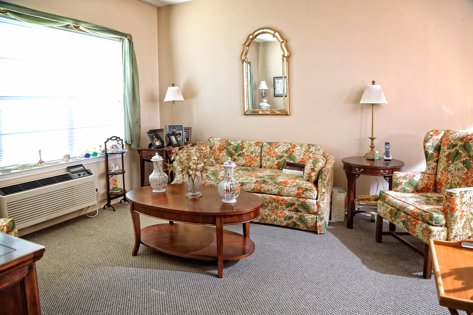 A cozy living room with floral-upholstered sofa and armchairs, a wooden coffee table, side tables with lamps, and a large window.