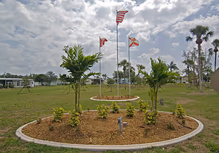 A landscaped circular garden bed with small palm trees and shrubs, three flagpoles with American, Canadian, and Florida state flags, and a grassy field with scattered trees and buildings in the background under a partly cloudy sky.