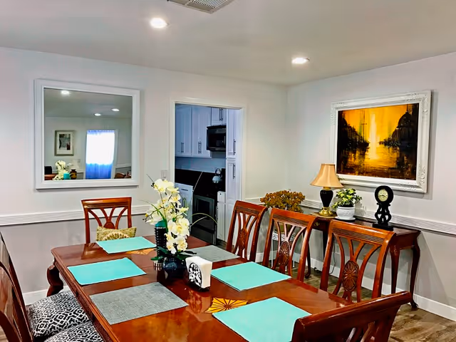 A dining room with a polished wooden table set with six chairs. The table has placemats in turquoise and gray, a vase with white flowers, and a tissue box. On the wall, there is a large mirror reflecting part of the room and a framed painting with warm yellow and black tones. A sideboard holds a lamp, a clock, and a small plant. The room has white walls and recessed ceiling lights.