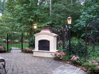 Outdoor patio area with a stone fireplace, surrounded by a paved stone walkway, black wrought iron fencing, lamp posts, and pink flowering plants, with dense green trees in the background.