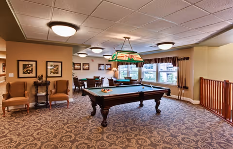 A spacious recreational room with a pool table in the center, surrounded by patterned carpet flooring. There are two upholstered chairs and a small table against one wall, framed artwork on the walls, and a large window with curtains letting in natural light. Ceiling lights and a hanging lamp above the pool table illuminate the room.