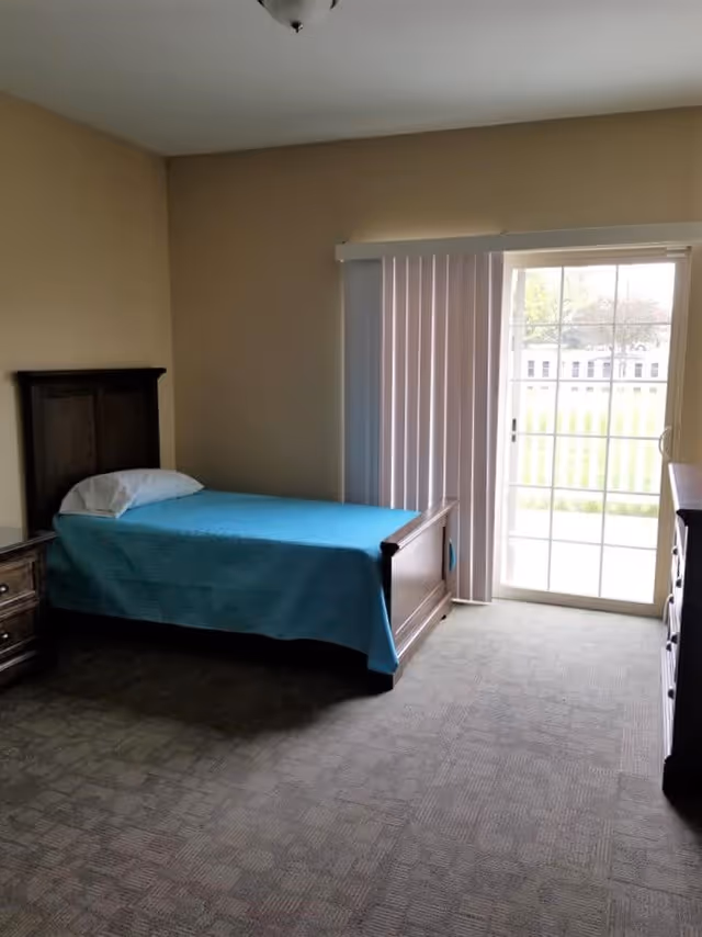 A simple bedroom with a single bed covered in a blue blanket, wooden furniture, carpeted floor, and a sliding glass door with vertical blinds.