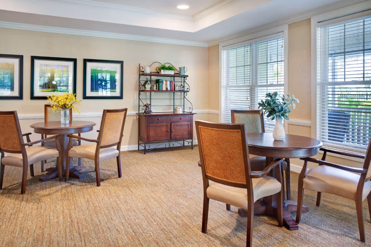 Bright communal dining area with round wooden tables and chairs, framed artwork, shelving and large windows.