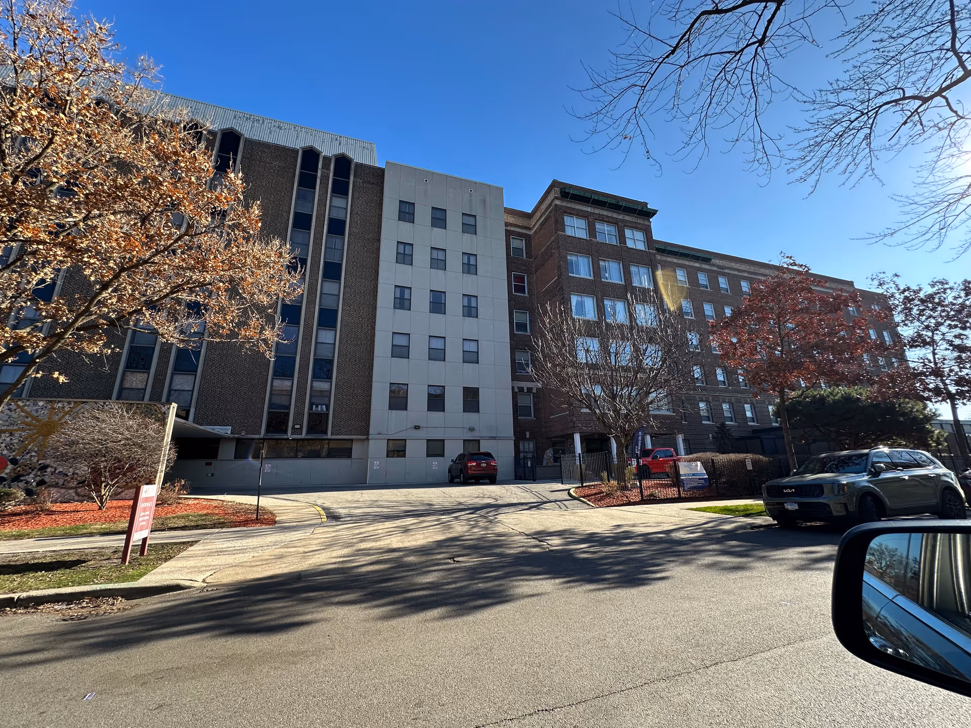Exterior view of a multi-story senior living facility building under a clear blue sky with some trees with autumn leaves and parked cars in front.