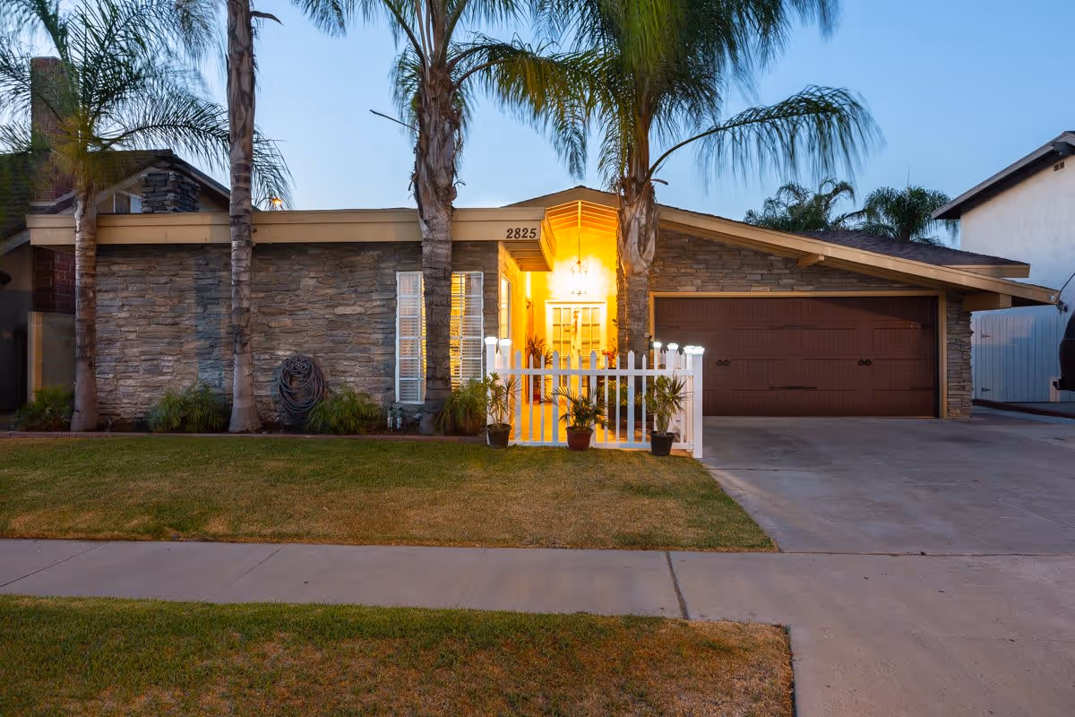 Front exterior view of a single-story house with stone facade, a brown garage door, three tall palm trees, a small white picket fence with potted plants, and a warmly lit entrance at dusk.