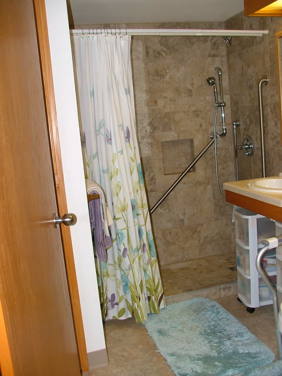 Accessible bathroom with a tiled walk-in shower behind a floral curtain, grab bars, sink and a small plastic storage cart.