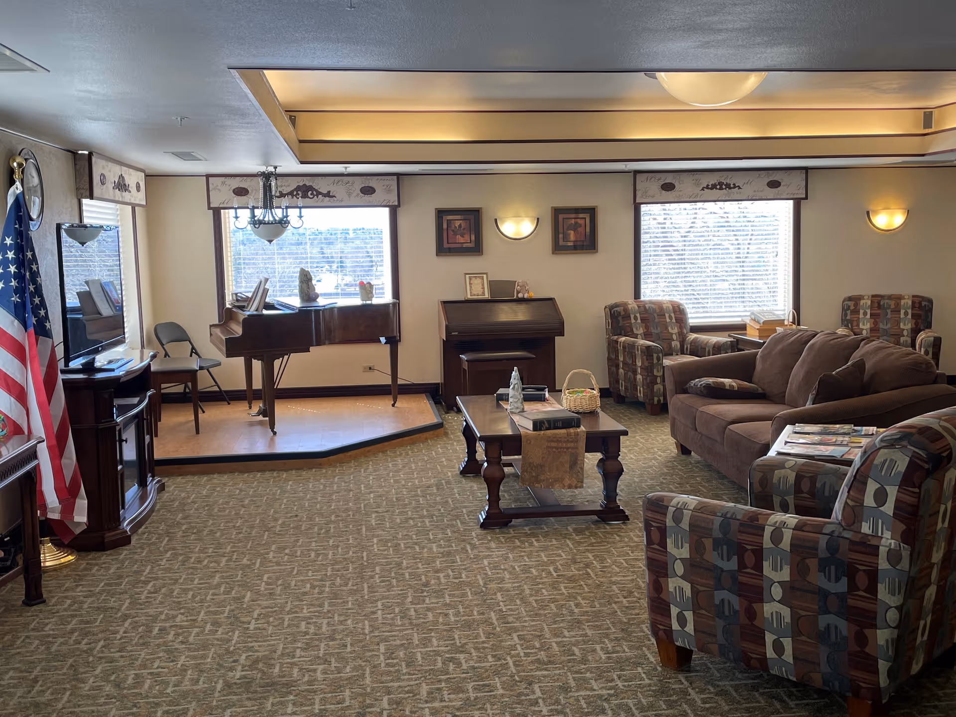 A cozy common room with sofas and patterned armchairs around a coffee table, a small stage with a grand piano, and an American flag near a TV.