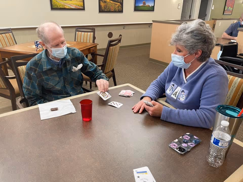 Two elderly individuals wearing face masks sitting at a table playing cards in a common area with wooden chairs and framed landscape pictures on the wall.