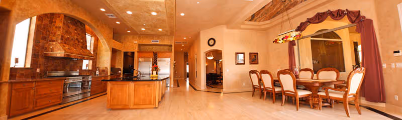 Spacious interior view of a senior living facility featuring a large kitchen area with wooden cabinets, a central island, and modern appliances on the left side. On the right side, there is a formal dining area with a wooden dining table and six upholstered chairs, illuminated by a decorative hanging light fixture. The walls have a warm beige tone with arched doorways and a clock mounted above one of the arches.